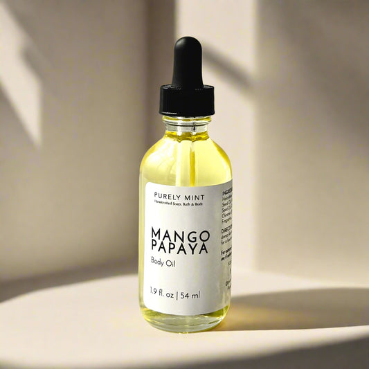 A 2 oz bottle of Mango Papaya Body Oil with a dropper top and a clean white label on a tan background. The sleek design showcases the minimalist aesthetic, with the amber-colored oil visible through the clear glass bottle.