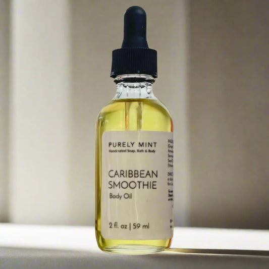 A 2 oz bottle of Caribbean Smoothie Body Oil with a dropper top and a clean white label on a tan background. The sleek design showcases the minimalist aesthetic, with the amber-colored oil visible through the clear glass bottle.