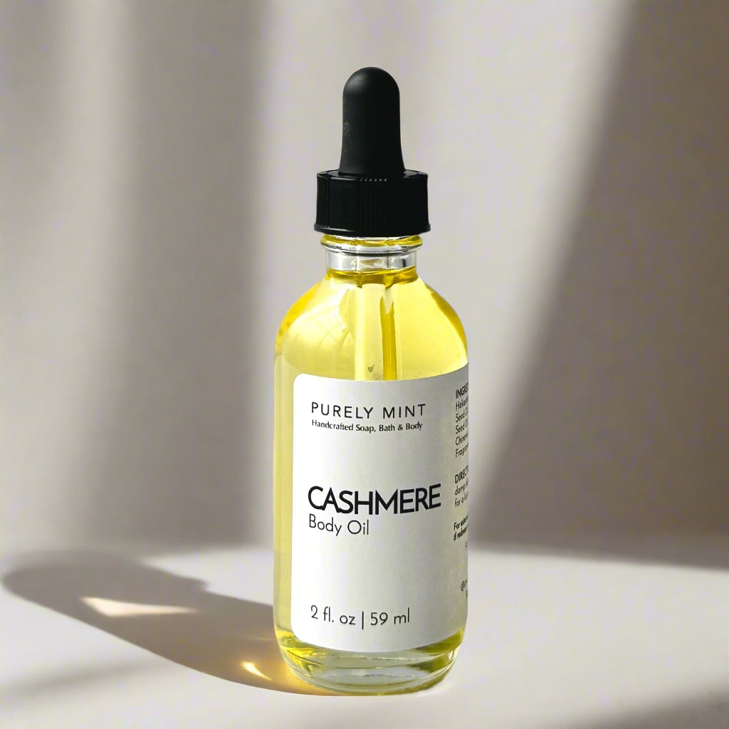A 2 oz bottle of Cashmere Body Oil with a dropper top and a clean white label on a tan background. The sleek design showcases the minimalist aesthetic, with the amber-colored oil visible through the clear glass bottle.