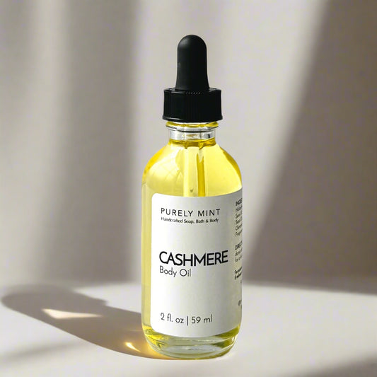 A 2 oz bottle of Cashmere Body Oil with a dropper top and a clean white label on a tan background. The sleek design showcases the minimalist aesthetic, with the amber-colored oil visible through the clear glass bottle.