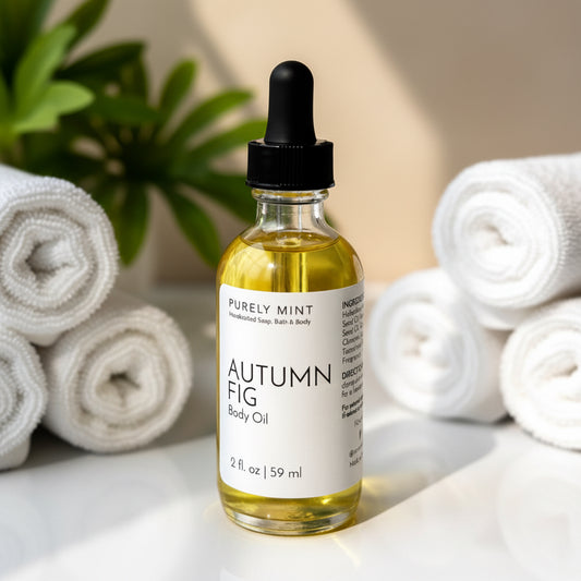A bottle of Autumn Fig body oil placed on a white counter, accompanied white rolled washcloths, green plant.
