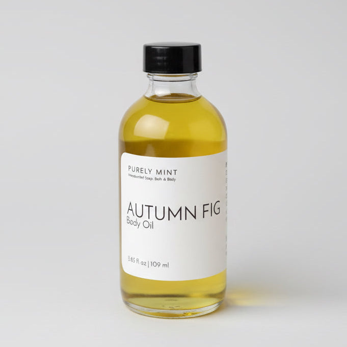 A 4 oz clear glass bottle of Autumn Fig body oil with a white label, placed on a light gray seamless backdrop, neutral lighting.