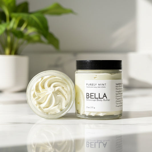 A open and closed jar of Bella body butter on a white marble counter with a green plant.