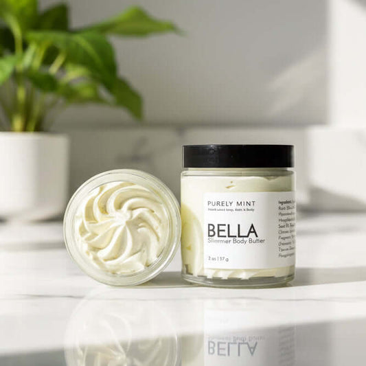 A open and closed jar of Bella shimmer body butter on a white marble counter with a green plant.