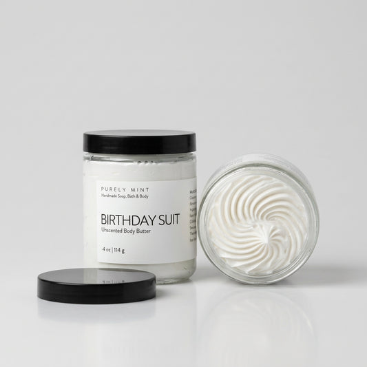 A glass jar of Purely Mint Birthday Suit unscented body butter with a black lid, standing next to a body butter showing the whipped cream texture inside, set on a light gray background.