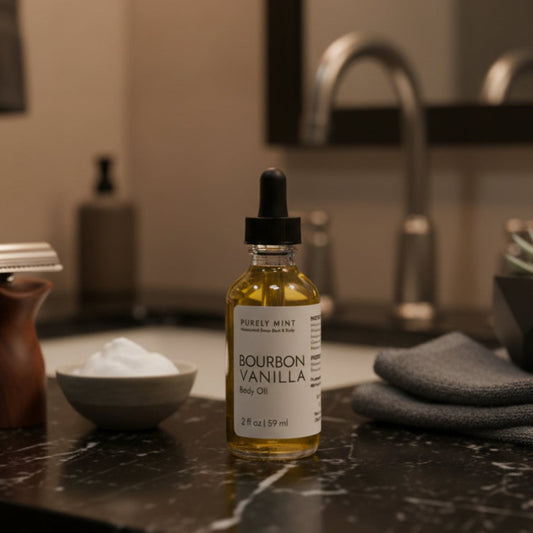 A bottle of Purely Mint Bourbon Vanilla Body Oil stands on a dark marble bathroom counter next to a razor, a small bowl of shaving cream, and a folded gray towel, with a blurred sink and mirror in the background.