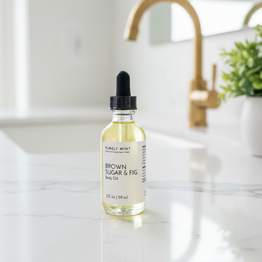 A bottle of Purely Mint Brown Sugar & Fig body Oil stands on a white marble countertop in a bright bathroom setting with a gold faucet and a potted plant in the background.