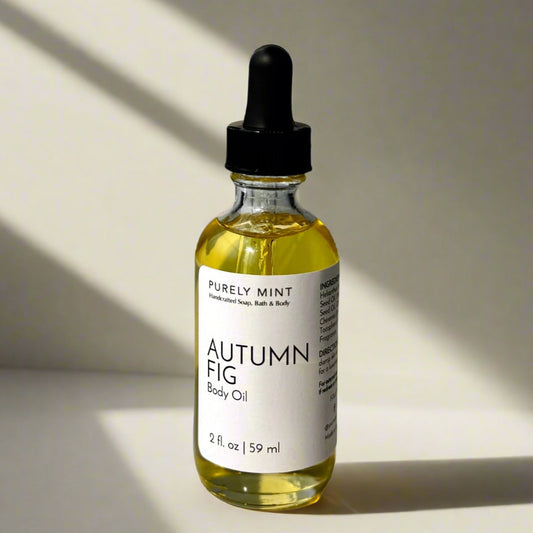 A 2 oz bottle of Autumn Fig Body Oil with a dropper top and a clean white label on a tan background. The sleek design showcases the minimalist aesthetic, with the amber-colored oil visible through the clear glass bottle.