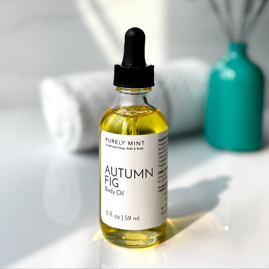 A bottle of Autumn Fig body oil placed on a white background, accompanied by a spa-style teal vase filled with green plants, creating a fresh and calming aesthetic.