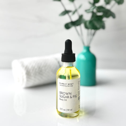 A bottle of Brown Sugar & Fig body oil placed on a white background, accompanied by a spa-style teal vase filled with green plants, creating a fresh and calming aesthetic.
