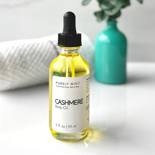 A bottle of Cashmere body oil placed on a white background, accompanied by a spa-style teal vase filled with green plants, creating a fresh and calming aesthetic.