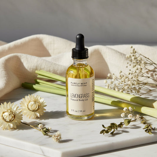 A bottle of Purely Mint Lemongrass Natural Body Oil stands on a white marble tray, surrounded by dried flowers, green stems, and draped cream fabric against a light gray background.