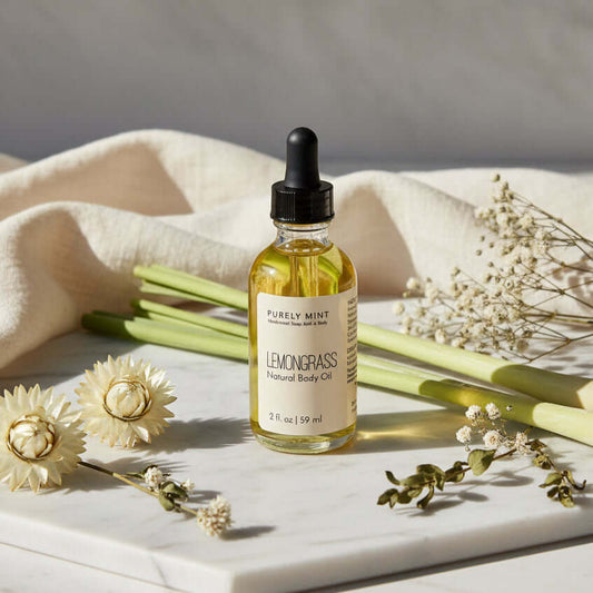 A bottle of Purely Mint Lemongrass Natural Body Oil stands on a white marble tray, surrounded by dried flowers, green stems, and draped cream fabric against a light gray background.