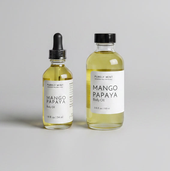 Two small glass bottles of Mango Papaya body oil with white labels and black dropper/cap closures, standing on a natural gray backdrop