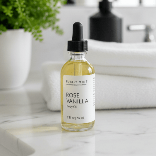 A bottle of Rose Vanilla Body Oil with a black dropper cap, set on a white marble bathroom counter with folded white towels, a potted plant, and a sink in the background.