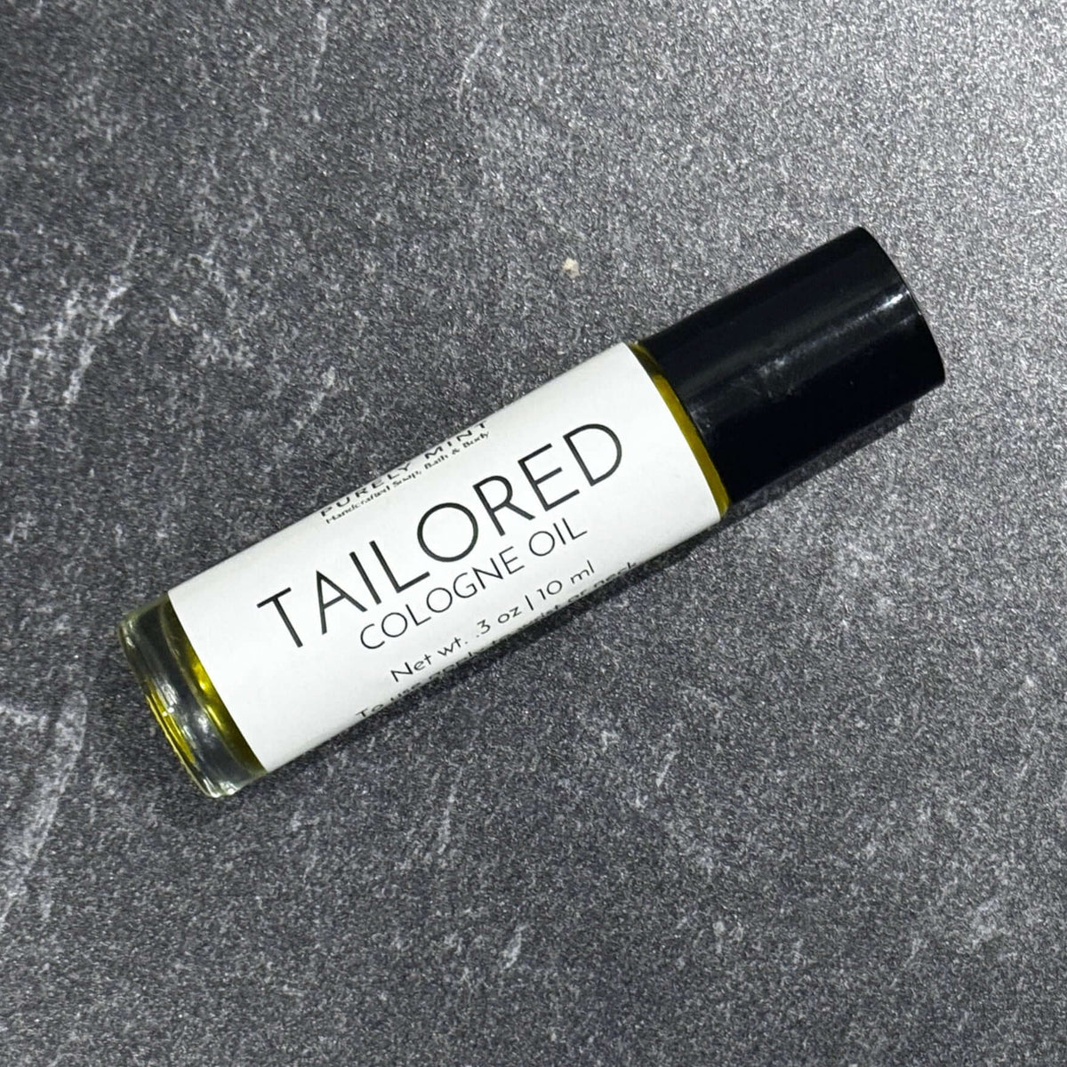 A 10 ml roll-on bottle of Tailored Cologne Oil by Purely Mint, featuring a white label and black cap, resting on a textured gray stone surface.