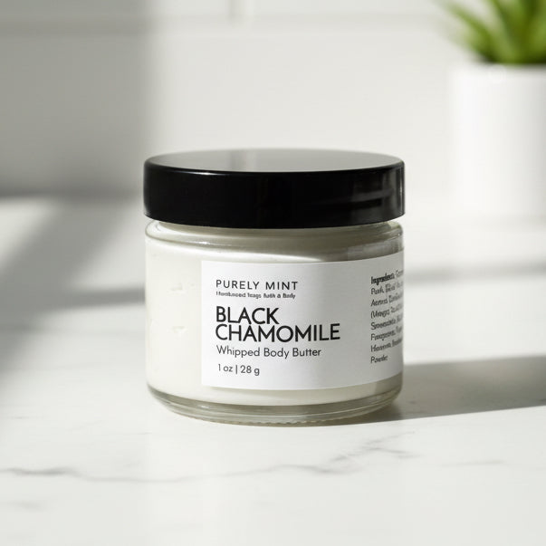 Black Chamomile whipped body butter jar in a modern bathroom, white marble counter, green plant.