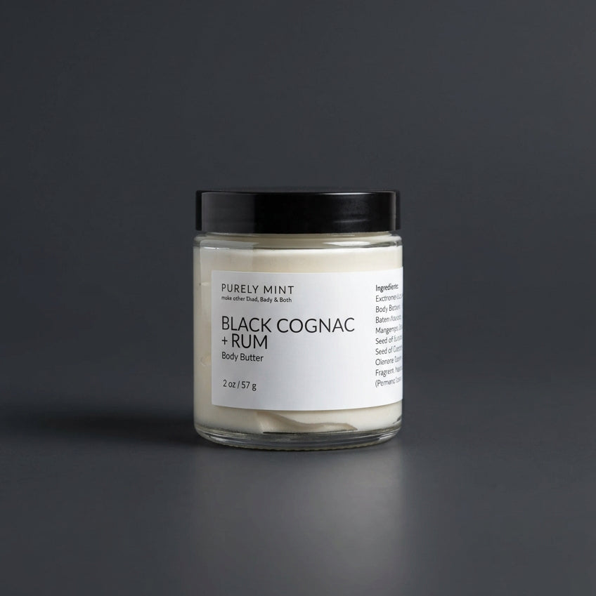 2 oz Black cognac & rum body butter in a clear glass jar with a white label and sleek black top on a dark gray surface.