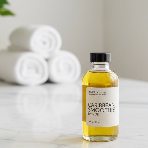 Bottle of Caribbean Smoothie body oil on a white marble counter with 3 wash cloths rolled and a green plant leaf