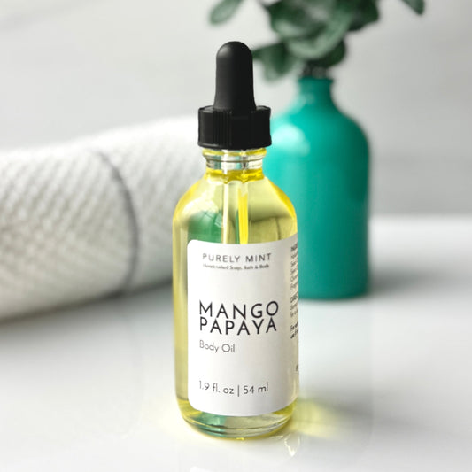 A bottle of 2 oz Mango Papaya body oil placed on a white background, accompanied by a spa-style teal vase filled with green plants, creating a fresh and calming aesthetic.