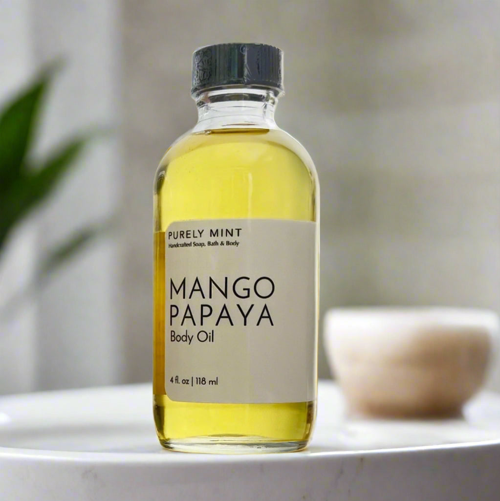 A 4 oz clear glass bottle of Mango Papaya body oil with a screw top and a simple white label. It is set against a minimalist, modern bathroom backdrop featuring neutral warm tones and spa-like elements, including green plants for a touch of natural luxury.