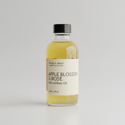 A 4 oz clear glass bottle of Apple Blossom and Rose body oil with a white label, placed on a light gray seamless backdrop.