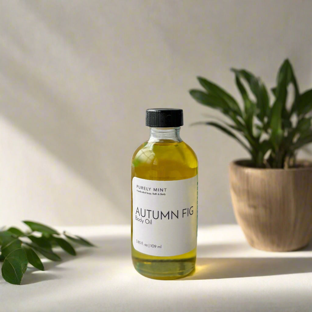 A 4 oz clear glass bottle of Autumn Fig body oil with a white label, placed in a softly sunlit setting. The scene features natural props including wooden elements, and green plants,  creating a minimalist aesthetic with neutral, warm tones. The lighting casts gentle shadows, highlighting the bottle's details and texture.