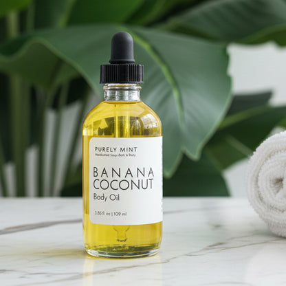 Bottle of banana coconut oil on a  white marble counter, green plant and rolled washcloth.