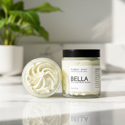 A open and closed jar of Bella body butter on a white marble counter with a green plant.