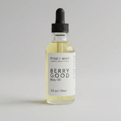 A bottle of Purely Mint Berry Good Body Oil with pale yellow oil inside, a white label, and a black cap, set against a plain bright gray backdrop background.