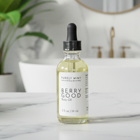 A bottle of Purely Mint Berry Good Body Oil with a black dropper cap and white label, set on a white marble countertop in a bright bathroom with a sink, gold faucet, and green plant in the background.