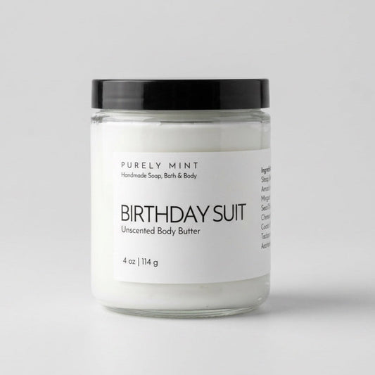 A glass jar containing white birthday suit unscented body butter rests on a light gray backdrop.