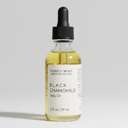 A 2 fl oz glass dropper bottle of Purely Mint Black Chamomile Body Oil, filled with pale yellow oil and featuring a white label and black dropper cap, set against a neutral gray background.