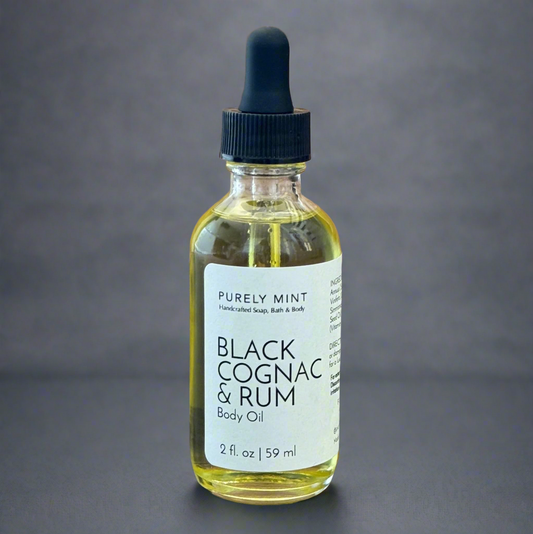 Black Cognac & Rum body oil, clear glass bottle, white label, black dropper top, commercial photography packshot, dark gray seamless backdrop.