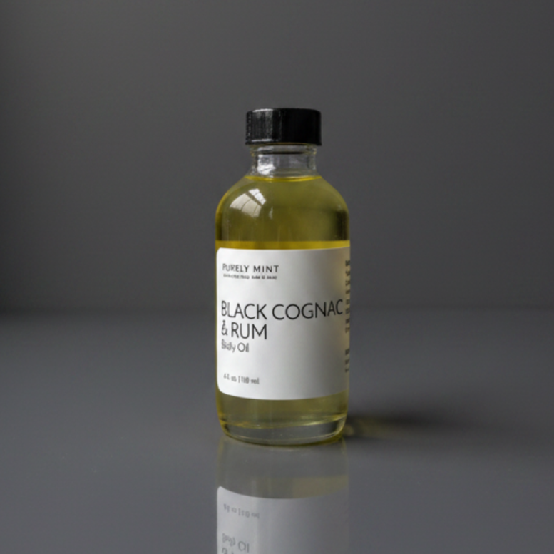 A bottle of Purely Mint Black Cognac & Rum Body Oil with pale yellow oil inside, a white label, a black cap, photographed on a dark gray reflective surface against a dark gray background.
