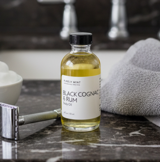A bottle of Purely Mint Black Cognac & Rum body oil stands on a dark marble bathroom counter next to a small bowl of cream, a metal razor, and a gray towel, with a blurred modern bathroom sink and mirror in the background.