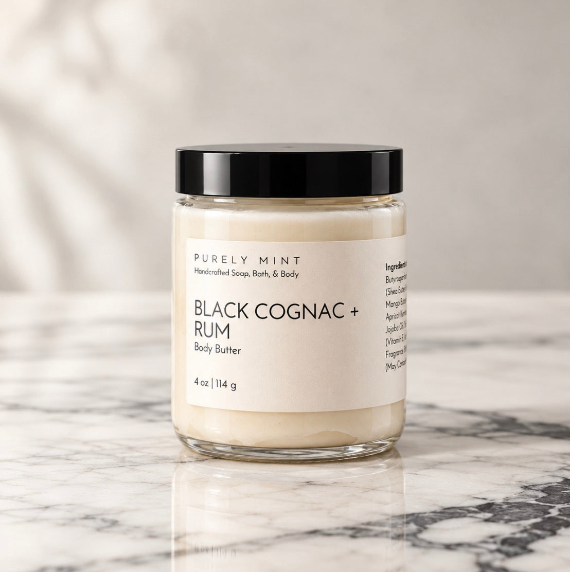 Black Cognac & Rum Body Butter by Purely Mint in a clear glass jar with a sleek black top on a marble surface