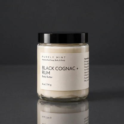 Black cognac & rum body butter in a clear glass jar with a white label and black sleek top on a reflective dark gray surface.