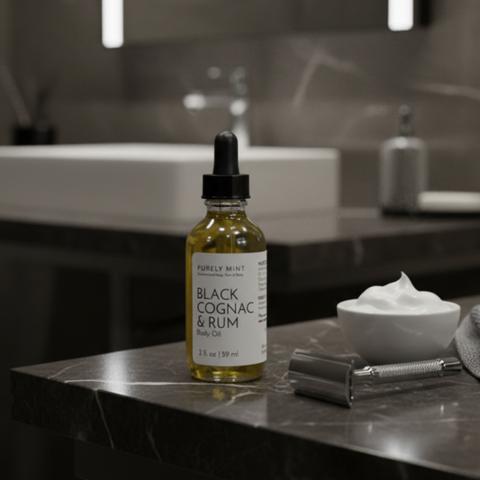 A bottle of Purely Mint Black Cognac & Rum body oil stands on a dark marble bathroom counter next to a small bowl of cream, a metal razor, and a gray towel, with a blurred modern bathroom sink and mirror in the background.
