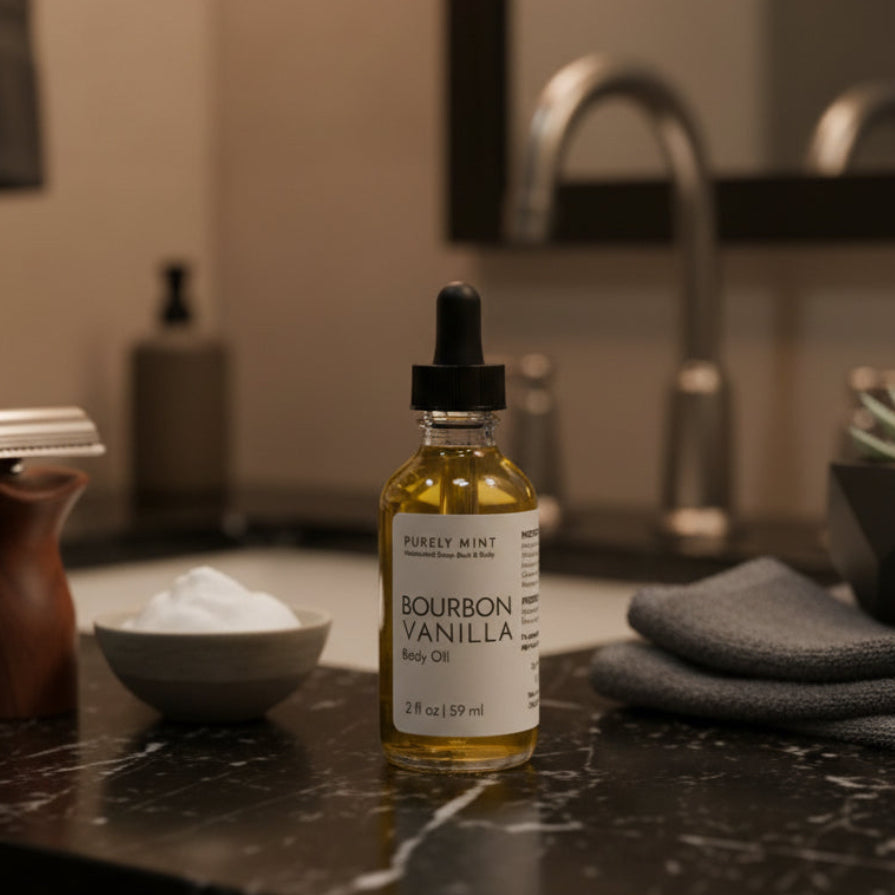 A bottle of Purely Mint Bourbon Vanilla Body Oil stands on a dark marble bathroom counter next to a razor, a small bowl of shaving cream, and a folded gray towel, with a blurred sink and mirror in the background.