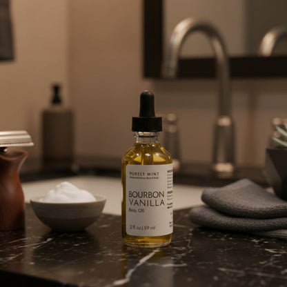 A bottle of Purely Mint Bourbon Vanilla Body Oil stands on a dark marble bathroom counter next to a razor, a small bowl of shaving cream, and a folded gray towel, with a blurred sink and mirror in the background.