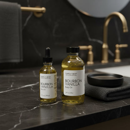 2 bottles of Purely Mint Body Bourbon Vanilla Body Oil with black caps are on a black marble bathroom counter next to a gray towel, and soap dish with a white sink and mirror in the background.