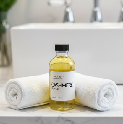 A bottle of Cashmere Body Oil by Purely Mint placed on a marble bathroom counter between two rolled white towels, with a white sink and faucet in the background.
