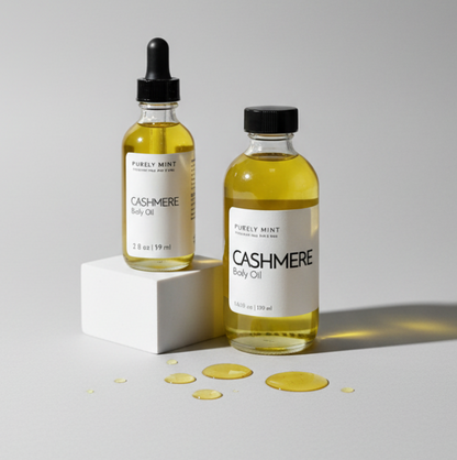 Two glass bottles of Purely Mint Cashmere Body Oil with yellow oil inside, one with a dropper cap and one with a black cap, displayed on a light gray background with a white block and oil droplets.