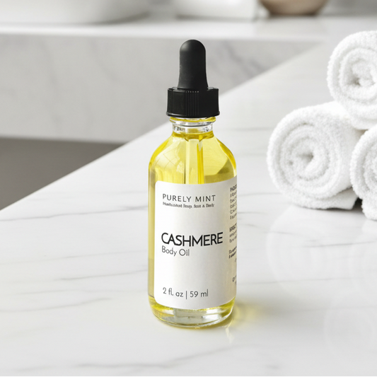 A bottle of Purely Mint Cashmere Body Oil with a black dropper cap sits on a white marble bathroom counter next to rolled white towels and a small potted plant.