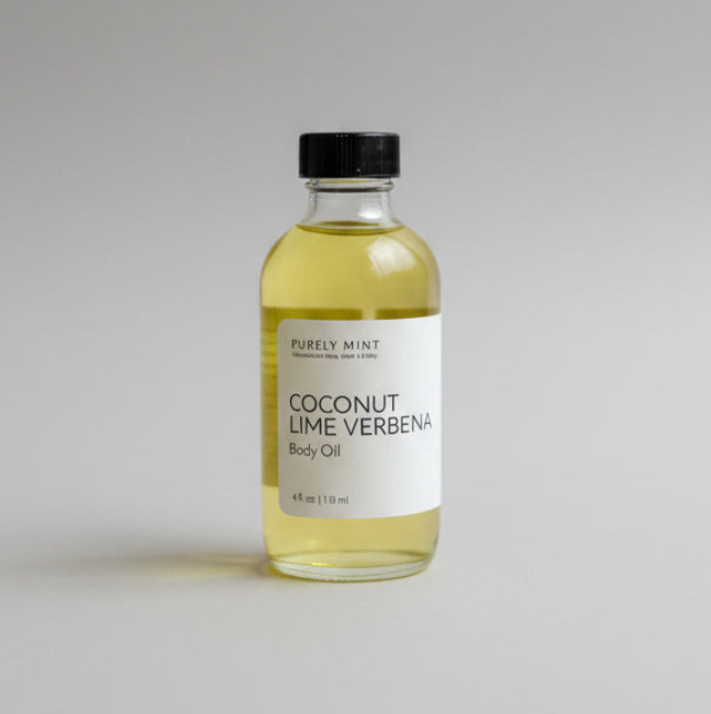 A clear glass bottle of Purely Mint Coconut Lime Verbena Body Oil with a black cap and white label, filled with pale yellow oil, set against a neutral light gray background.