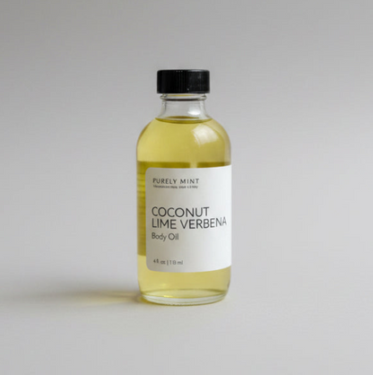 A clear glass bottle of Purely Mint Coconut Lime Verbena Body Oil with a black cap and white label, filled with pale yellow oil, set against a neutral light gray background.