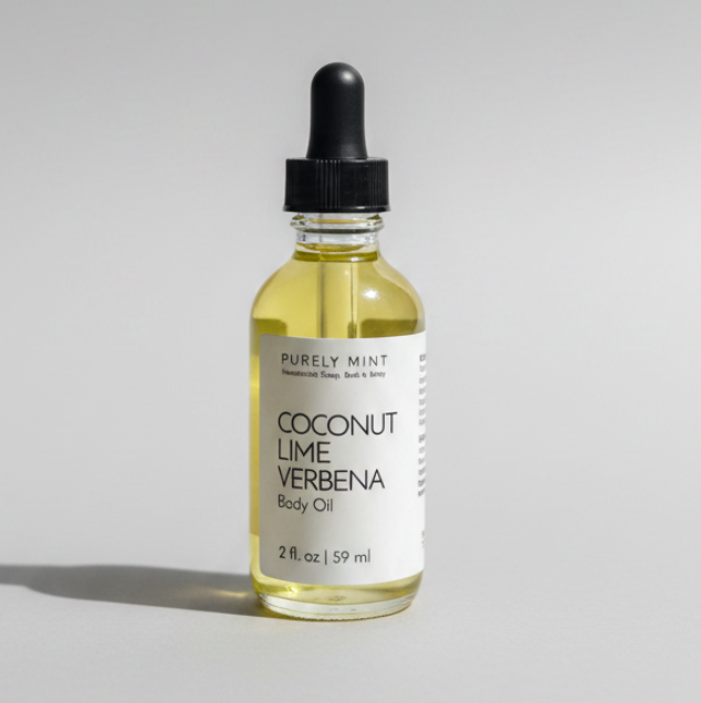 A clear glass bottle of Purely Mint Coconut Lime Verbena Body Oil with a black cap and white label, filled with pale yellow oil, set against a neutral light gray background.