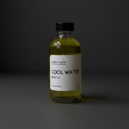 A bottle of Purely Mint 'Cool Water' body oil, featuring a clear glass bottle filled with yellow-green oil, a white label with black text, and a black cap, set against a dark gray background.
