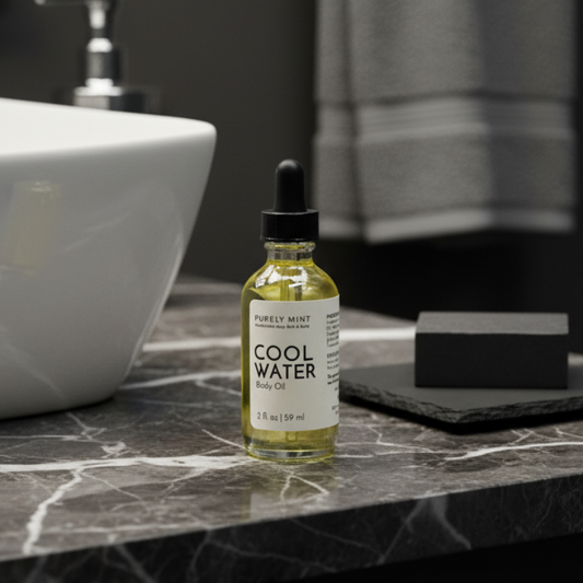 A 2 fl oz glass dropper bottle of Purely Mint Cool Water Body Oil, filled with pale yellow oil, standing on a dark marble counter, with a white sink and a soap dish with a black soap on it and a gray towel in the background.